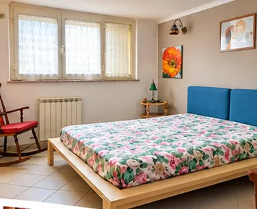Camera matrimoniale con letto in legno, testiera imbottita blu, finestra luminosa e poltrona a dondolo