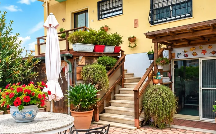 Terrazza esterna della casa vacanze a Ladispoli, con tavolo in pietra, sedie in ferro battuto, ombrellone e piante in vaso davanti all'ingresso